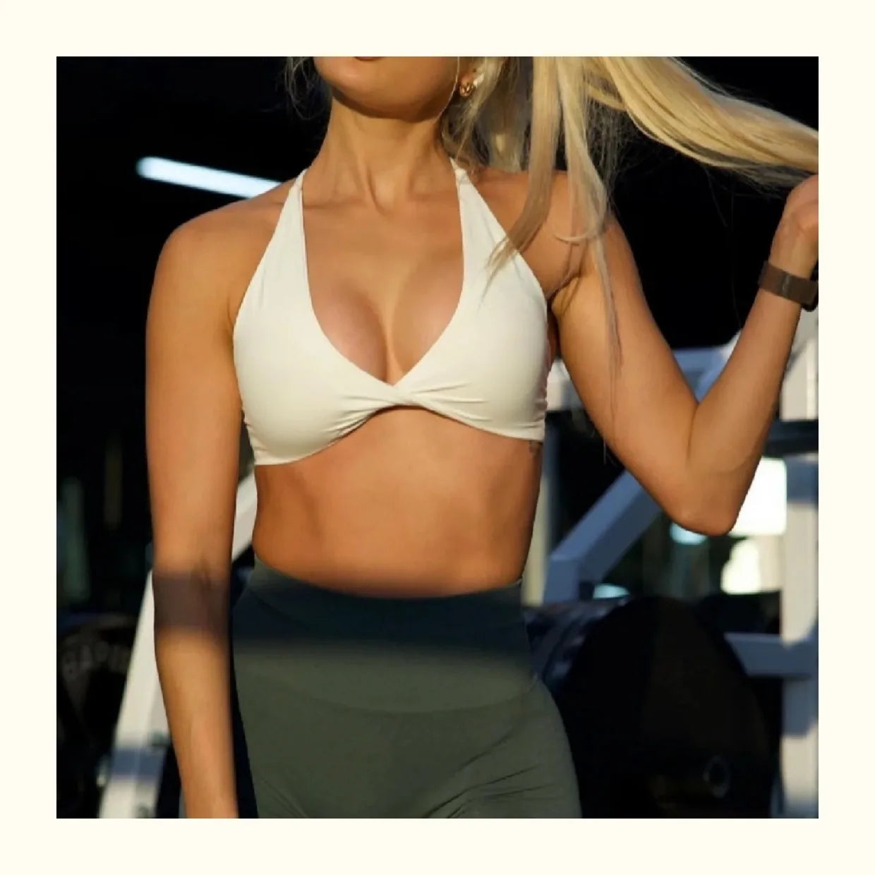 Woman wearing white padded front twist sports bra and dark green leggings in gym setting