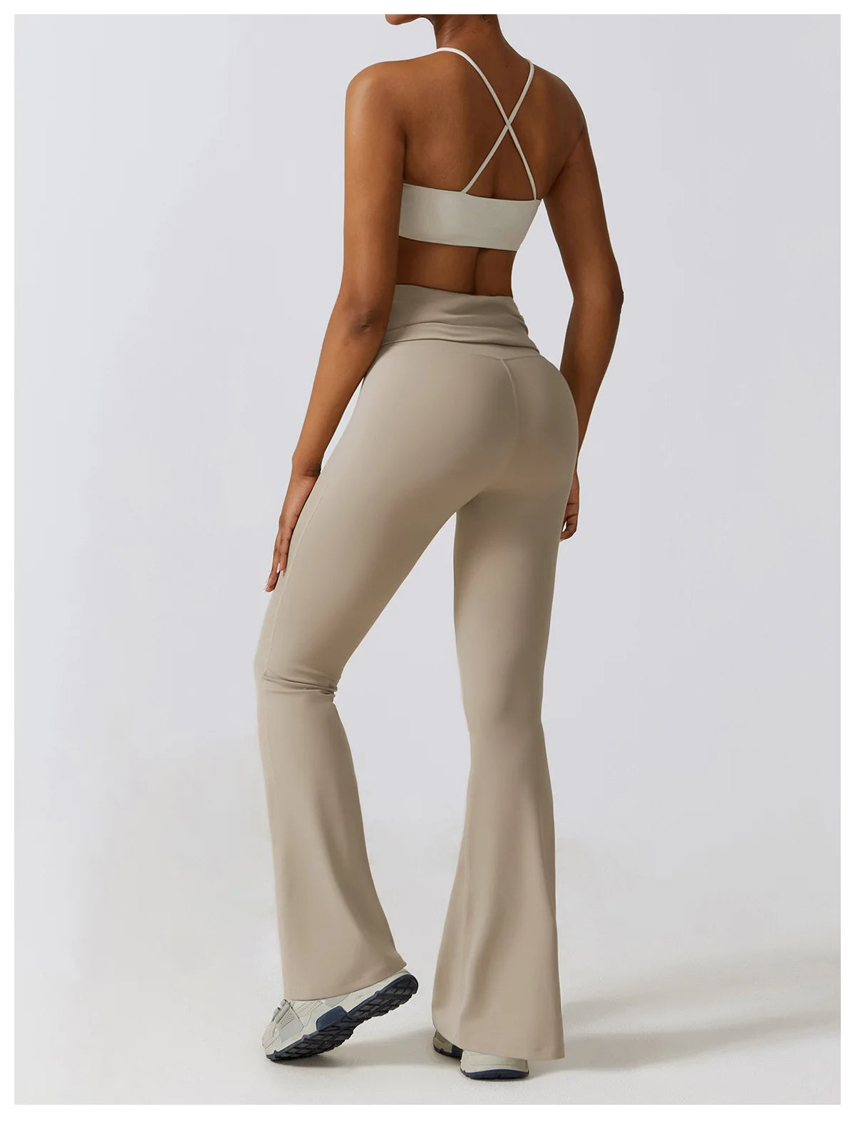 Back view of a woman wearing beige one piece yoga suit for women with flared pants and cross strap top
