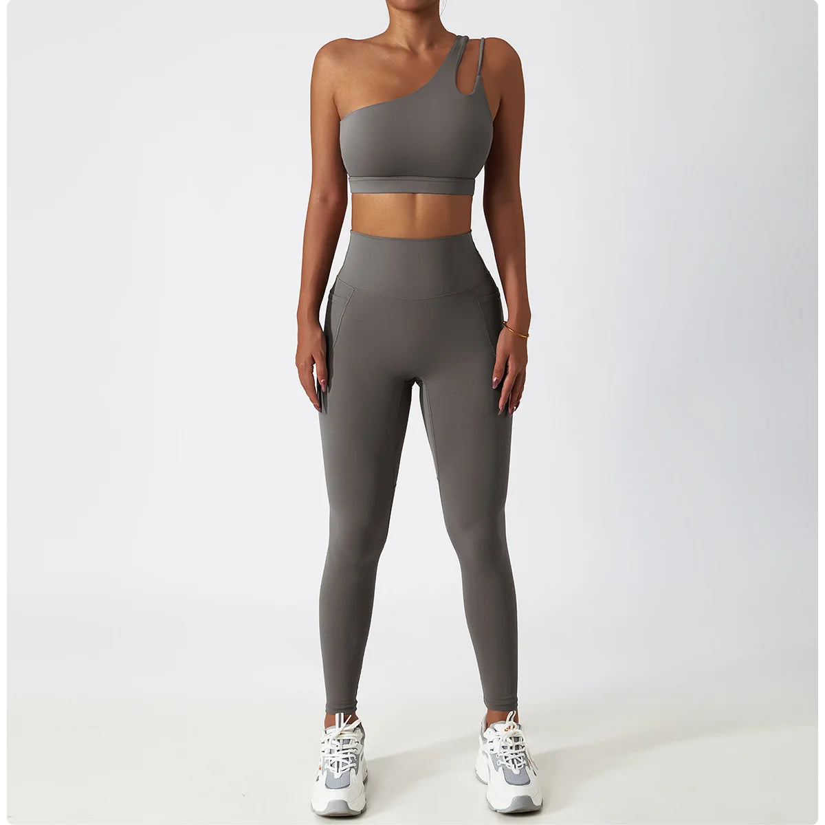 KiKis one shoulder sports bra and leggings set in gray modeled with white sneakers on white background