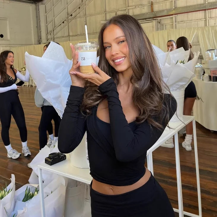 Smiling woman wearing a black women's long sleeve yoga top holding a cold coffee drink indoors