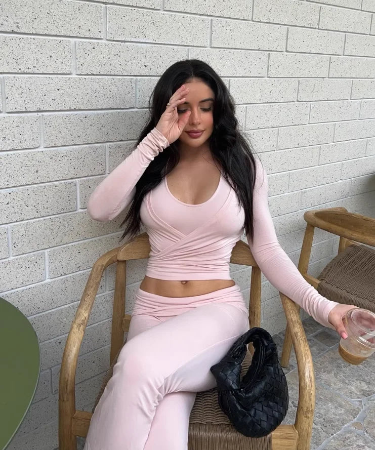 KiKis women's long sleeve yoga top in light pink worn with matching pants seated on wooden chair