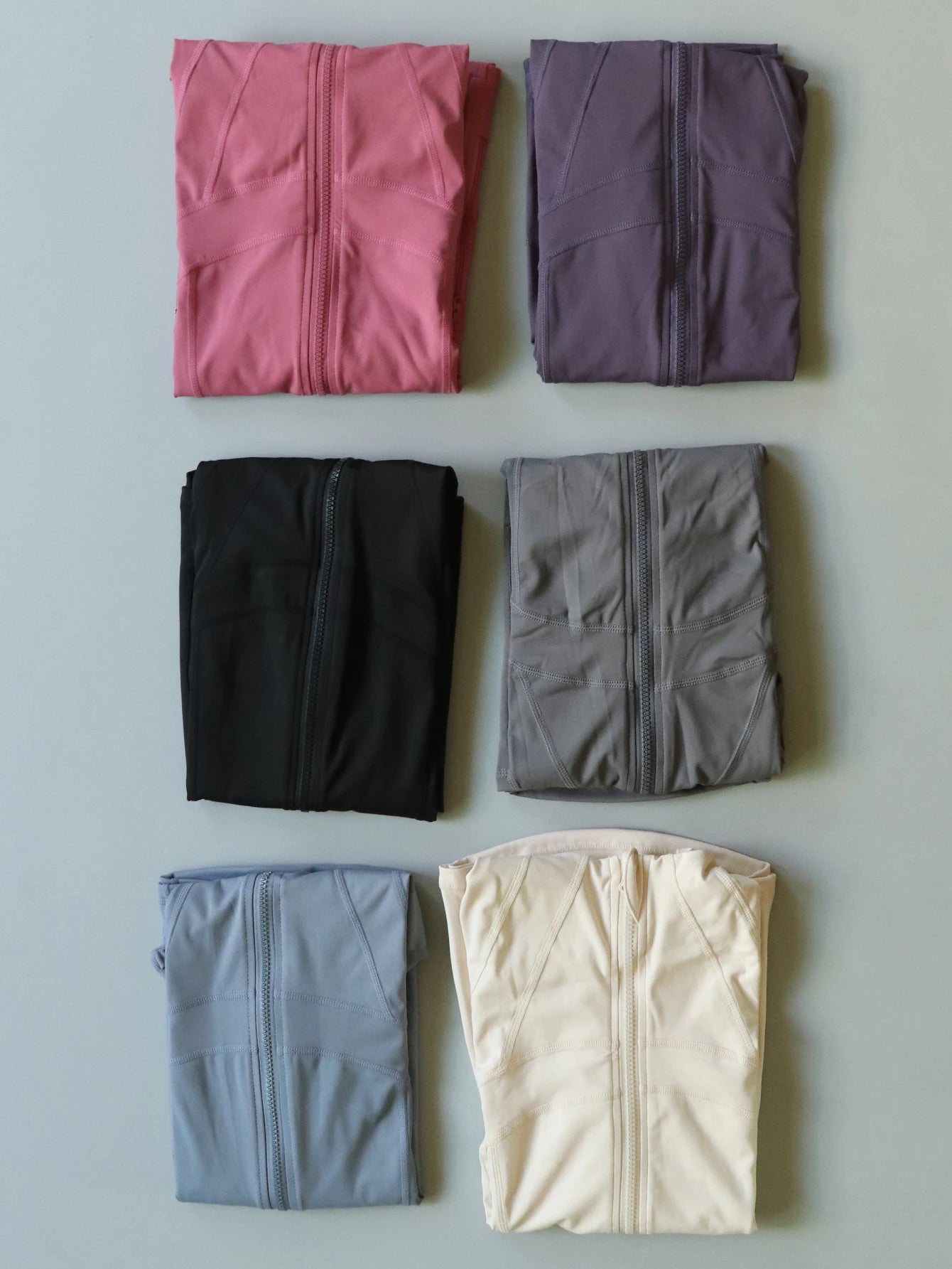 Six folded Women's Lightweight ZipUp Activewear Jackets in pink, purple, black, gray, blue, and cream colors neatly arranged