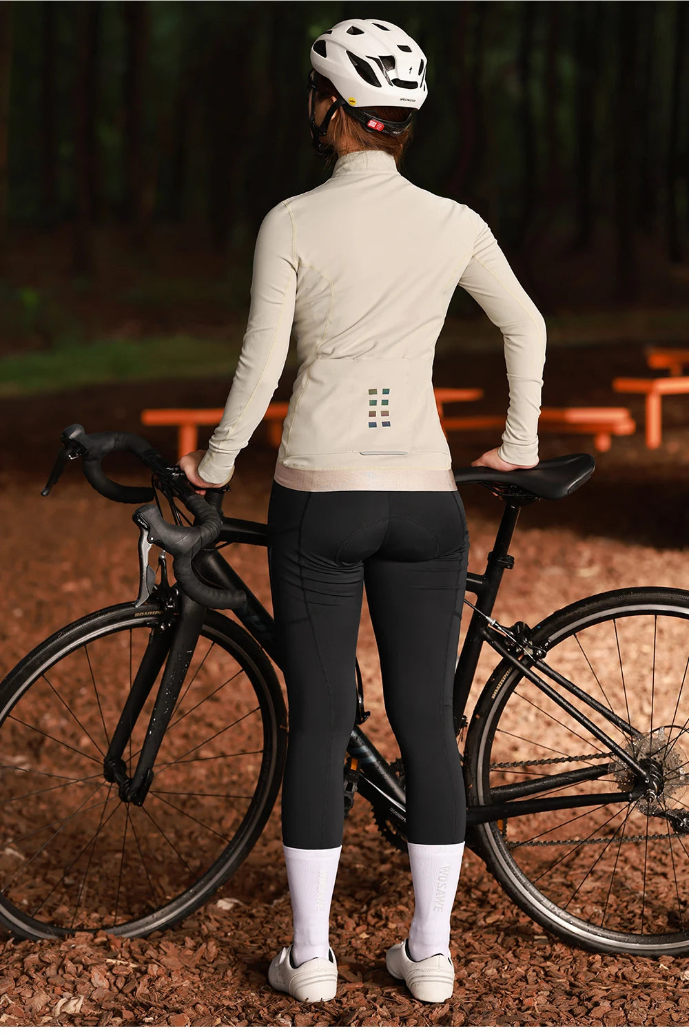 Woman wearing KiKis women's winter cycling jacket standing next to a black road bike in a forest setting